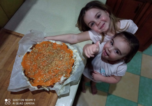 Dwie dziewczynki pozują do zdjęcia wraz z ciastem ustawionym na blacie.